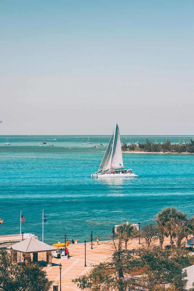 A WeRoad group trip sails on a white catamaran across turquoise water, viewed from a waterfront plaza under a clear sky.