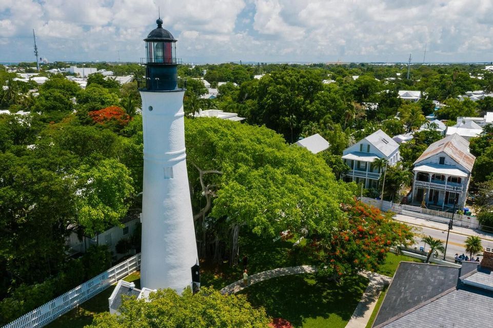 An aerial view of a tall, white lighthouse with a black top standing among lush green trees and houses in a coastal town.