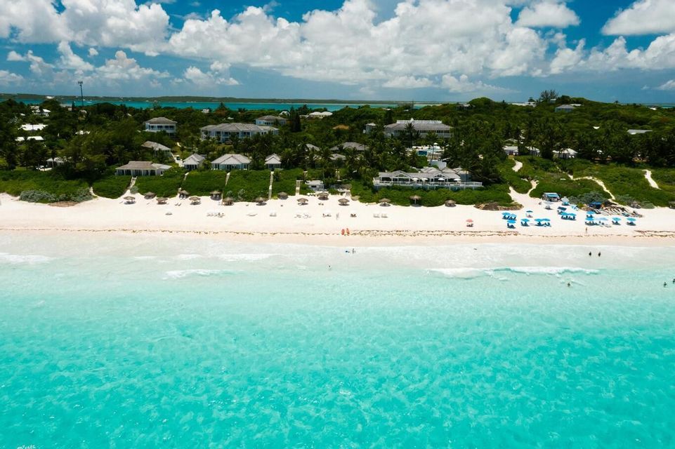 An aerial view of a tropical beach with white sand, clear turquoise water, and resorts nestled in lush green foliage under a partly cloudy sky.