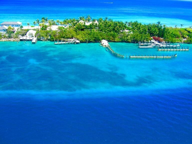 An aerial view of a small tropical island with palm trees and docks, surrounded by turquoise and deep blue ocean water.
