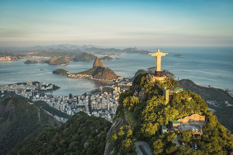 An aerial view of the Christ the Redeemer statue on a mountain overlooking a city, a bay with islands, and Sugarloaf Mountain.
