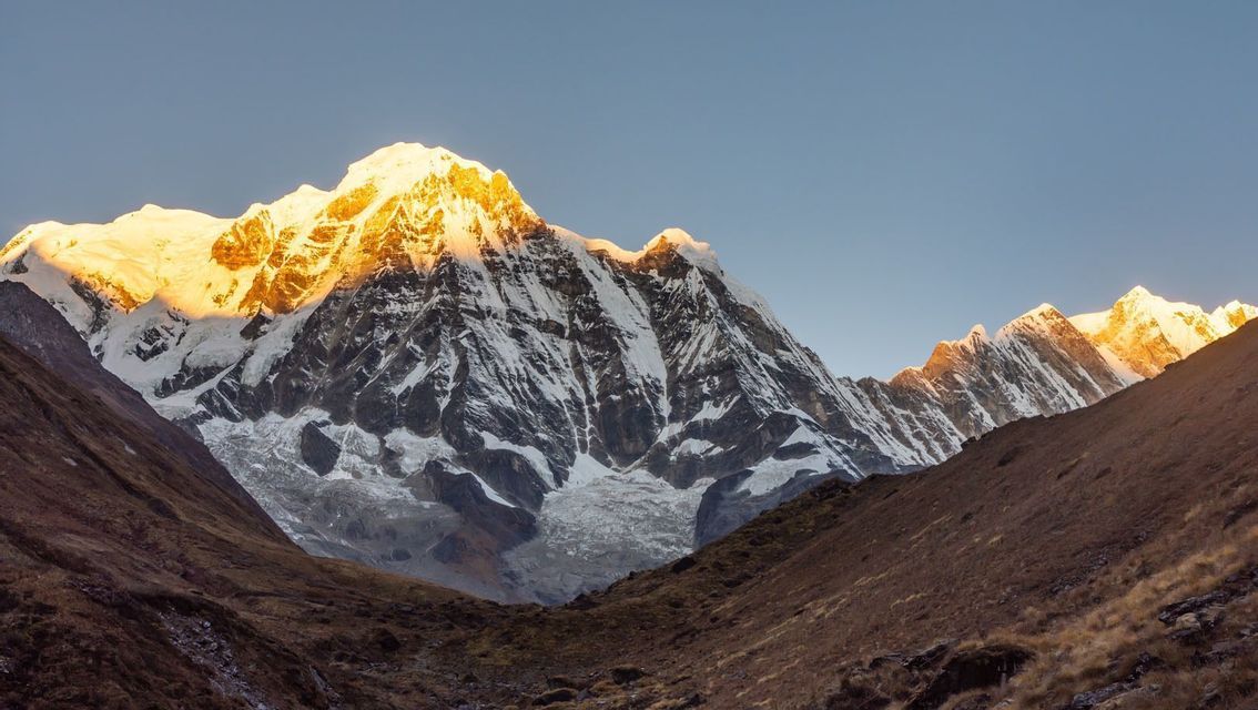 La lumière dorée du lever ou du coucher de soleil illumine les sommets enneigés d'une chaîne de montagnes, vus d'une vallée rocheuse en contrebas.