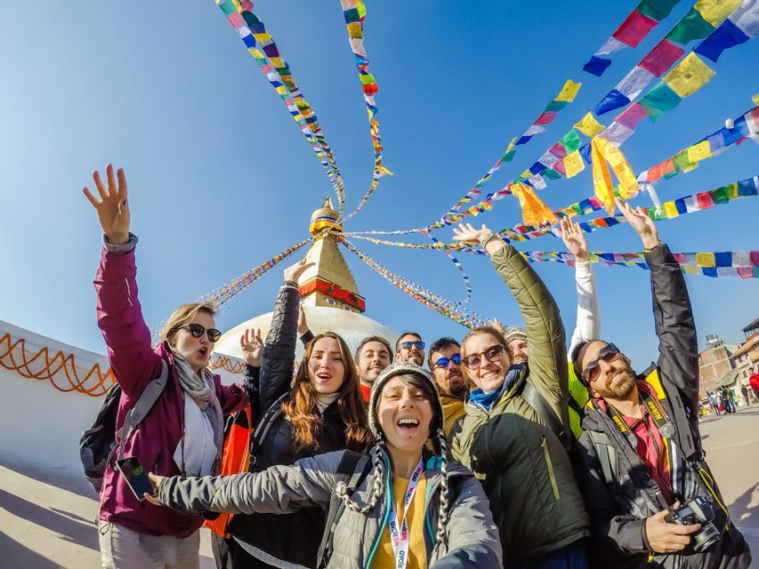 Un viaggio di gruppo WeRoad scatta un selfie sorridente davanti a una grande stupa sotto bandiere di preghiera colorate e un cielo blu limpido.