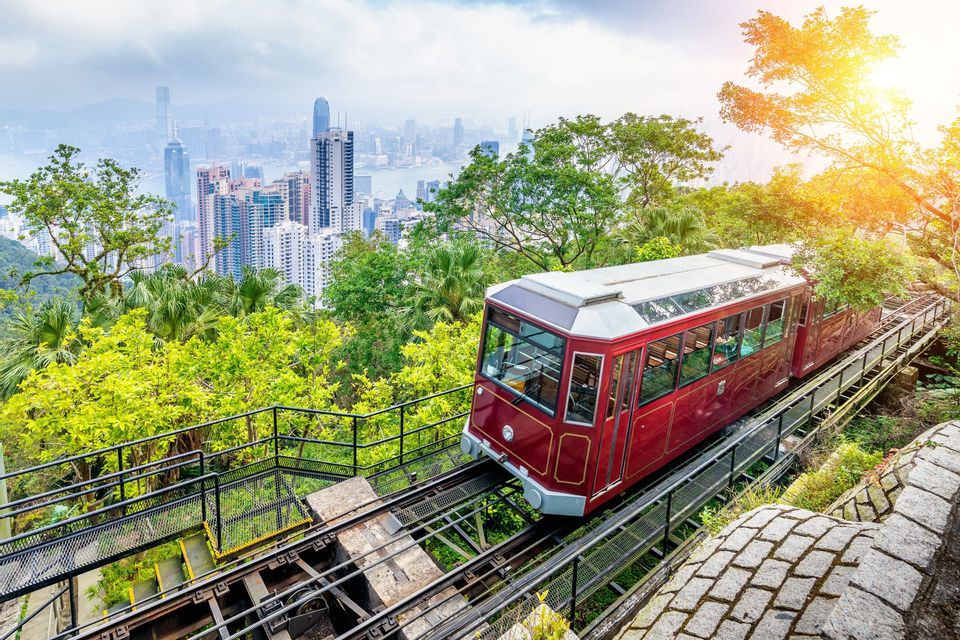 Un funicular rojo asciende por una vía en una exuberante ladera verde, con una vista panorámica de una moderna ciudad al fondo.