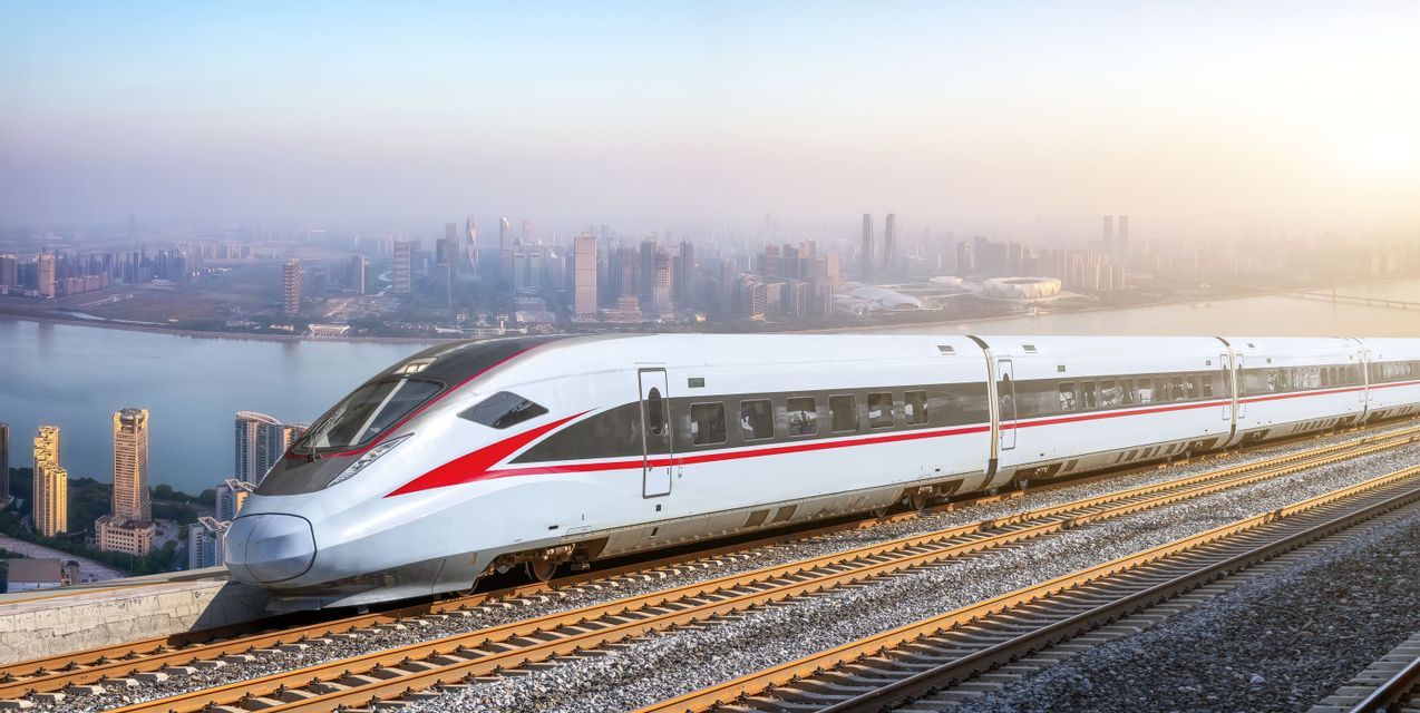 Un tren blanco de alta velocidad con una franja roja viaja por las vías, con vistas al horizonte de una ciudad moderna al otro lado del agua al amanecer.