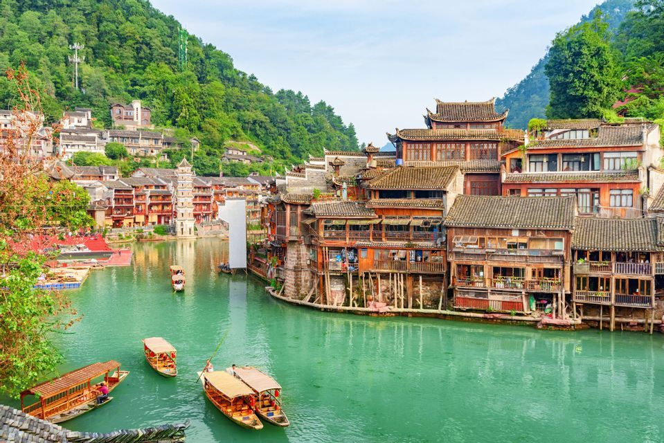 Un pueblo antiguo con casas tradicionales de madera sobre pilotes y botes en un río turquesa, rodeado de exuberantes montañas verdes.