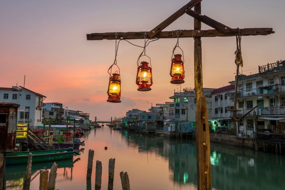 Tres farolillos rojos iluminados cuelgan sobre un canal en un pueblo palafítico al atardecer, con barcas amarradas a lo largo del agua.