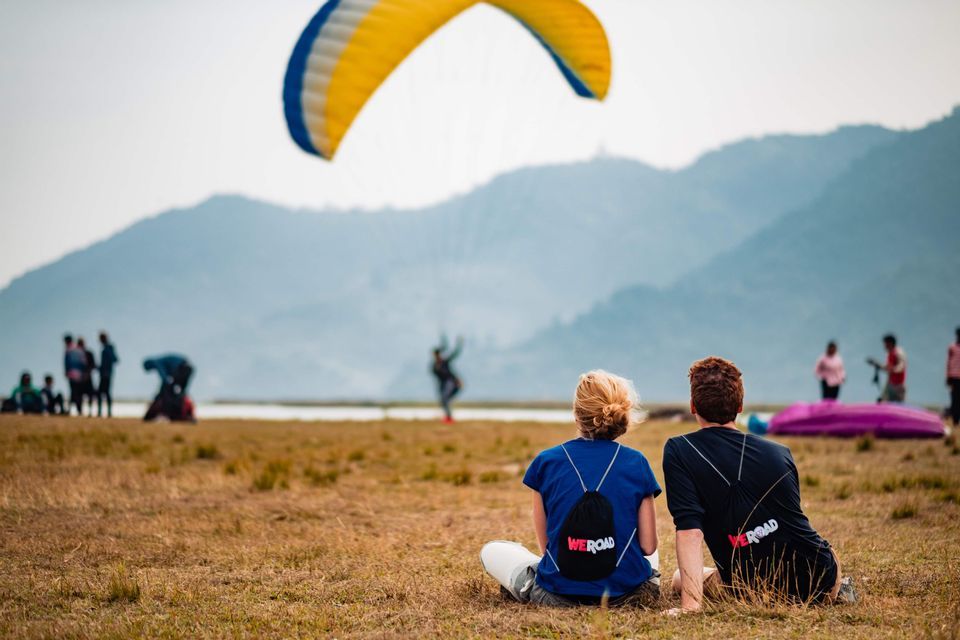 Two people from a WeRoad group trip sit on a grassy field watching a yellow and blue paraglider fly over distant mountains.