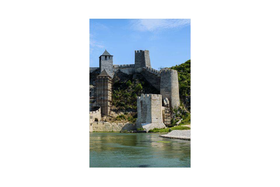 Una fortezza medievale in pietra con più torri si erge su un verde pendio, al margine di un ampio fiume, sotto un cielo blu.