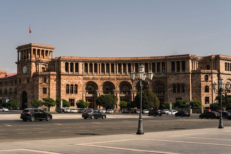 Un bâtiment monumental en pierre avec une tour d'horloge et des colonnades arquées se dresse sur une vaste place de ville avec des voitures.