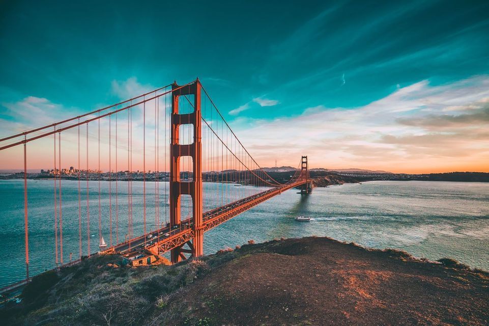 Die Golden Gate Bridge von einem Hügel bei Sonnenuntergang aus gesehen, mit einer Stadtsilhouette in der Ferne über dem Wasser.