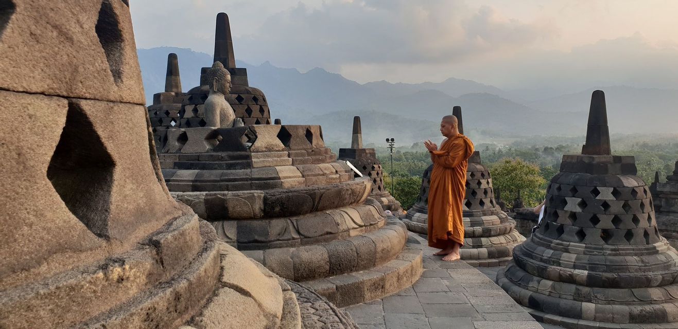 A monk in an orange robe prays barefoot among stone stupas at an ancient temple, with mountains in the background.