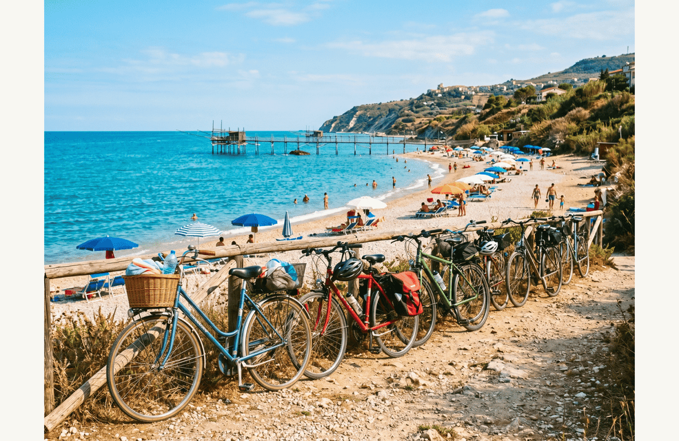 Una fila di biciclette parcheggiate su un sentiero costiero si affaccia su una spiaggia sabbiosa con acqua turchese e un pontile di legno.