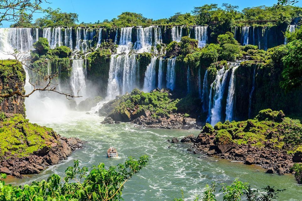 A small tourist boat navigates the turbulent river at the base of a large, multi-tiered waterfall, covered in lush green vegetation.