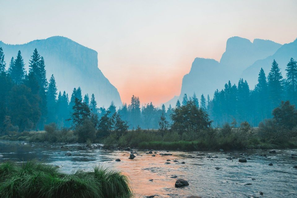 A rocky river flows through a pine forest with misty mountains in the background under a sunset sky.