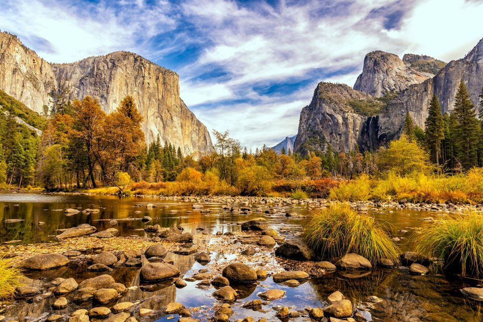 A wide valley with a rocky river, autumn foliage, and towering granite mountains under a partly cloudy sky.