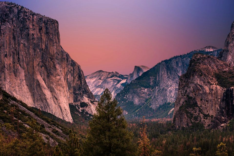 A vast mountain valley filled with a pine forest, flanked by steep granite cliffs under a purple and pink sky at dusk.