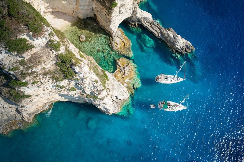 Luftbild von zwei Segelbooten, die in türkisblauem Wasser neben weißen Felsklippen und einer einsamen Bucht ankern.