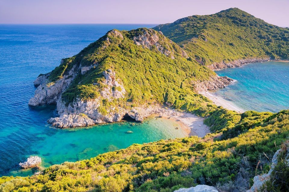 Ein Panoramablick auf eine üppig grüne Landzunge mit felsigen Klippen, die auf das türkisfarbene Meer treffen und einen abgelegenen Sandstrand in einer Bucht bilden.