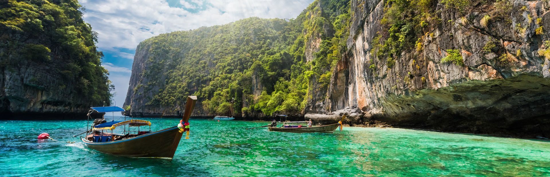 Des pirogues traditionnelles flottent dans un lagon turquoise, entouré de hautes falaises verdoyantes, sous un ciel partiellement nuageux.