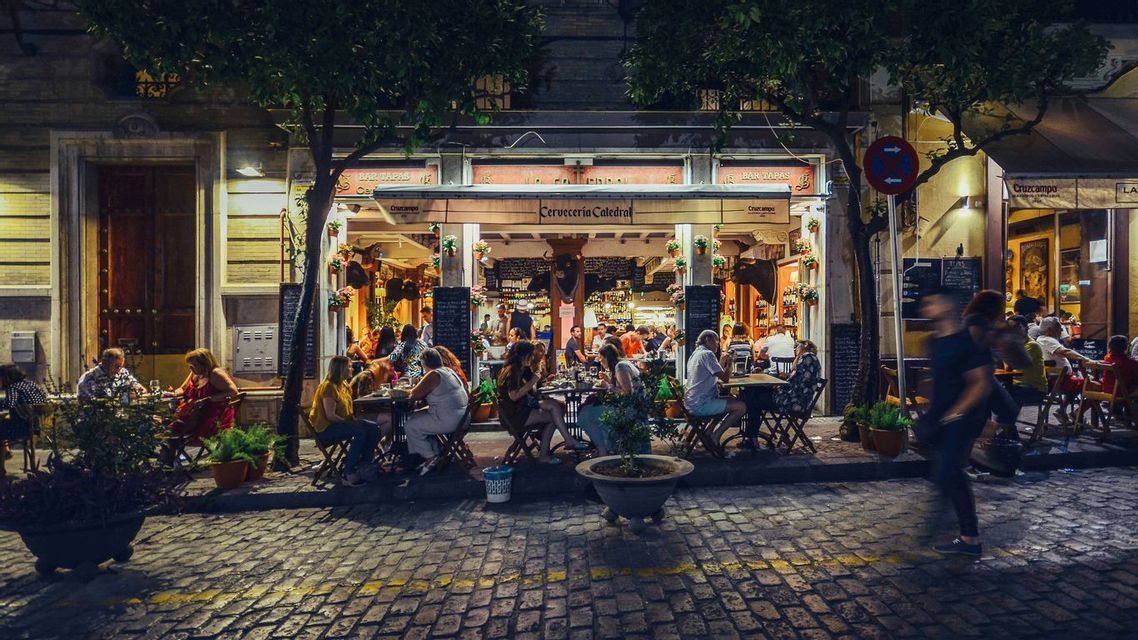 A bustling outdoor restaurant scene at night, with patrons dining at tables along a cobblestone street.