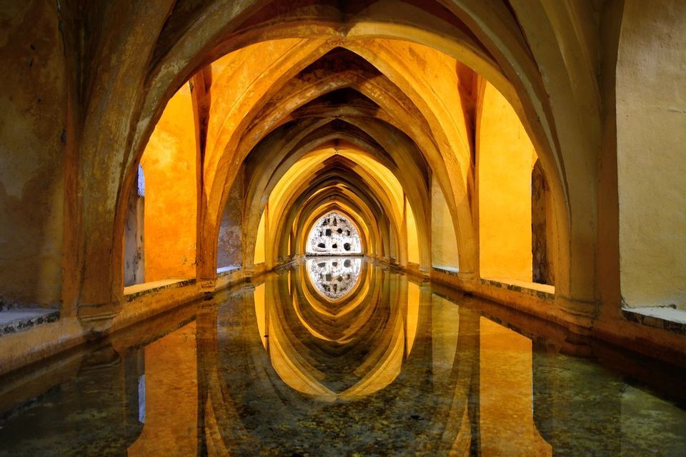 A long, arched hallway illuminated by golden light is reflected symmetrically in a narrow pool of still water.