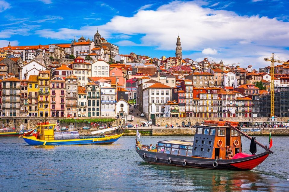 Two traditional boats sail on a river in front of a hillside city with colorful buildings under a partly cloudy blue sky.