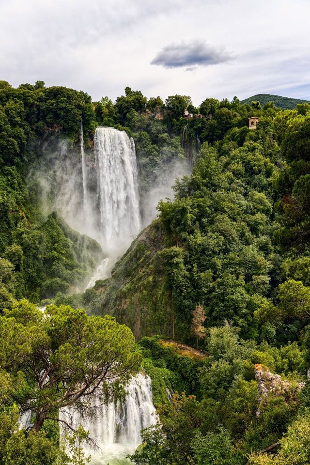 Una grande cascata scende da una scogliera rocciosa in una foresta lussureggiante e verde sotto un cielo nuvoloso.