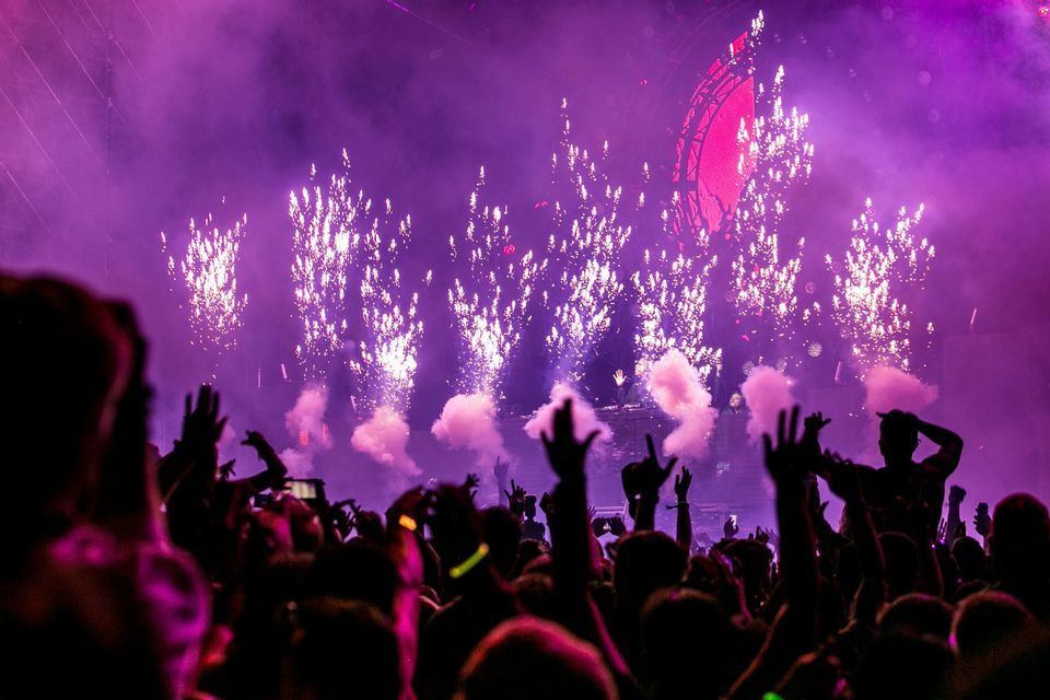 Des feux d'artifice explosent au-dessus d'une foule en silhouette lors d'un festival de musique avec des lumières violettes.