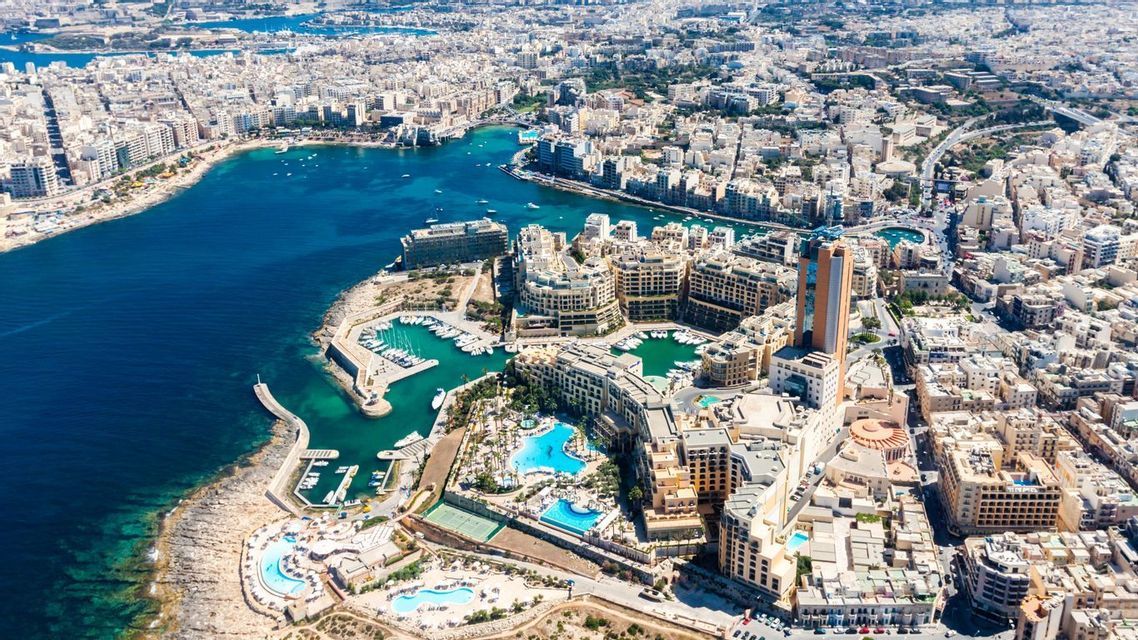 Une vue aérienne d'une ville côtière dense avec des bâtiments modernes entourant une marina remplie de bateaux par une journée ensoleillée.