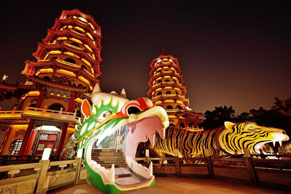 Deux grandes pagodes brillamment illuminées se découpent sur un ciel nocturne, avec d'imposantes statues colorées d'un dragon et d'un tigre au premier plan.