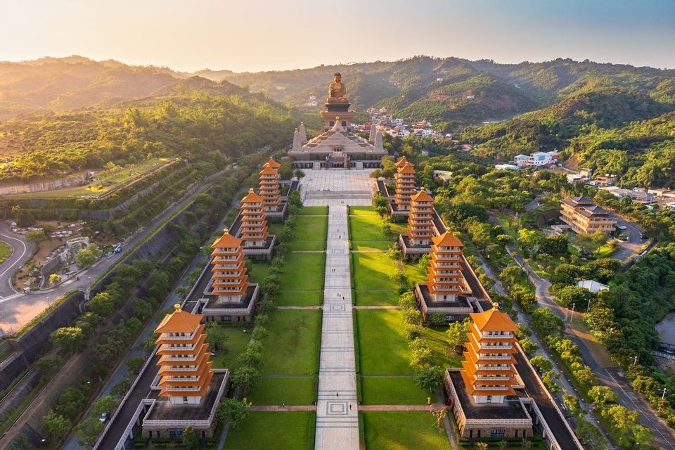 Vue aérienne d'un complexe de temples avec des pagodes bordant un chemin central menant à une grande statue de Bouddha, sur fond de collines verdoyantes.