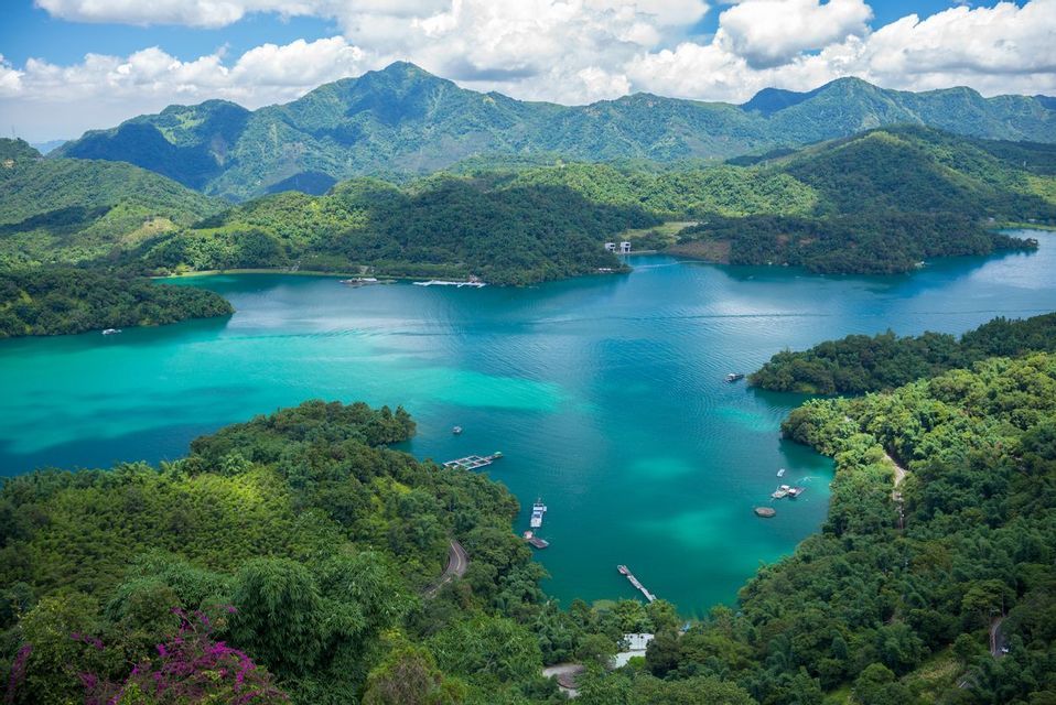 Vue aérienne d'un lac turquoise éclatant niché au cœur de montagnes verdoyantes, avec de petits bateaux parsemant l'eau sous un ciel nuageux.