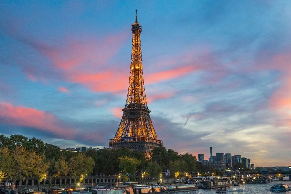 La Torre Eiffel illuminata si erge contro un cielo colorato al tramonto, con un fiume e lo skyline della città in primo piano.