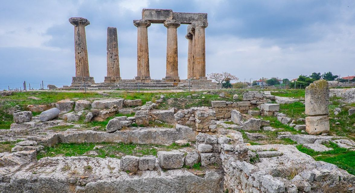 Alte colonne di pietra di un antico tempio si ergono tra le rovine su un campo erboso sotto un cielo nuvoloso.