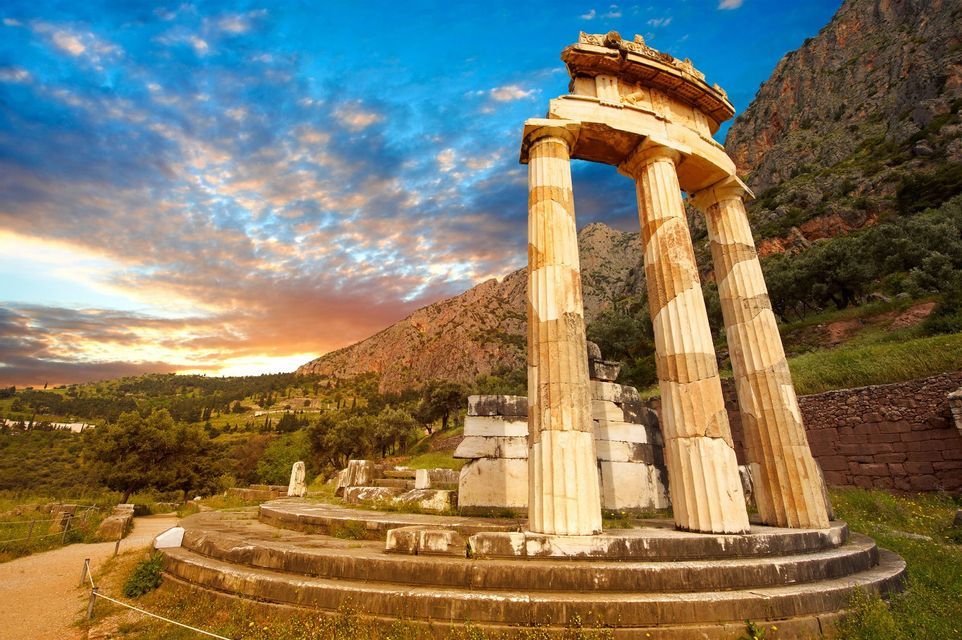 Le rovine di un antico tempio circolare con colonne sorgono su una base di pietra in una verde valle montuosa al tramonto.