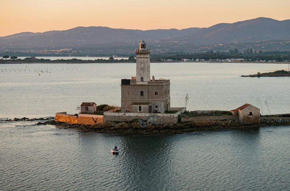 Un faro storico sorge su una piccola isola rocciosa in mezzo al mare, con una barchetta che galleggia nelle vicinanze sotto un cielo infuocato dal tramonto.