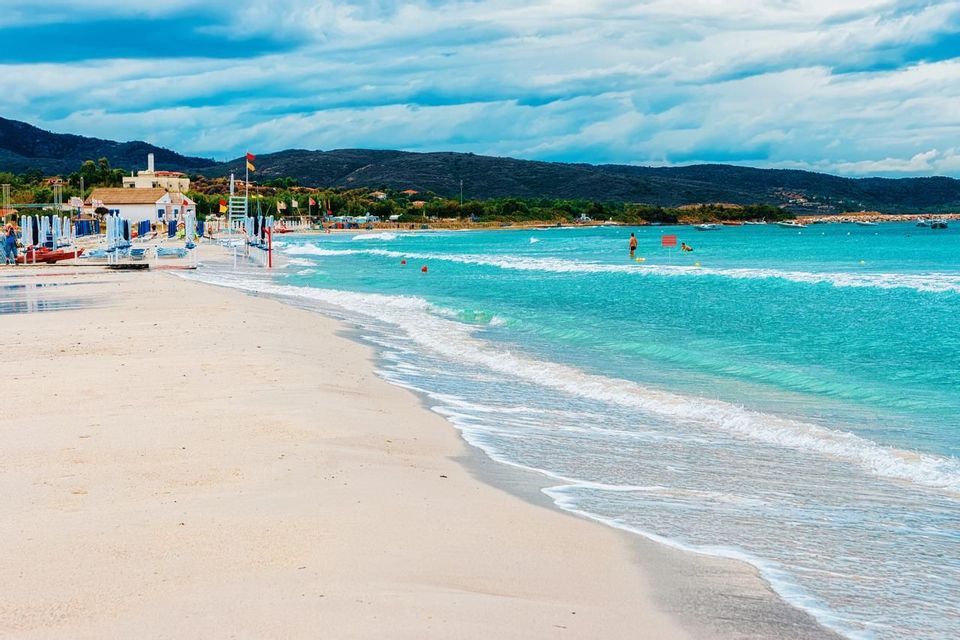 Una spiaggia di sabbia incontra l'oceano turchese, con piccole onde che lambiscono la riva. Sullo sfondo si scorgono lettini, edifici e colline.