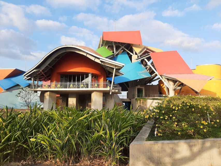 Un bâtiment coloré à l'architecture moderne et abstraite, doté de toits superposés et multicolores, est vu depuis un jardin.