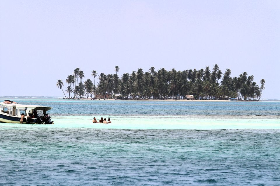 Un groupe WeRoad se baigne dans les eaux peu profondes et turquoise d'un banc de sable, avec leur bateau amarré à proximité et une île couverte de palmiers en arrière-plan.