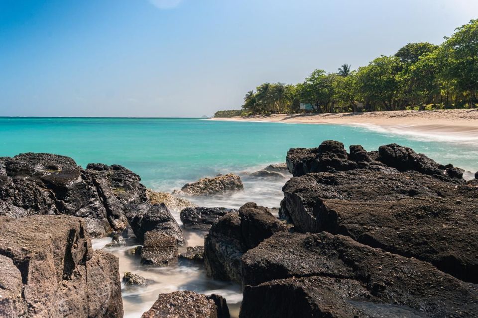 Lunga esposizione di acqua turchese che scorre su rocce scure su una spiaggia tropicale sotto un cielo azzurro chiaro.