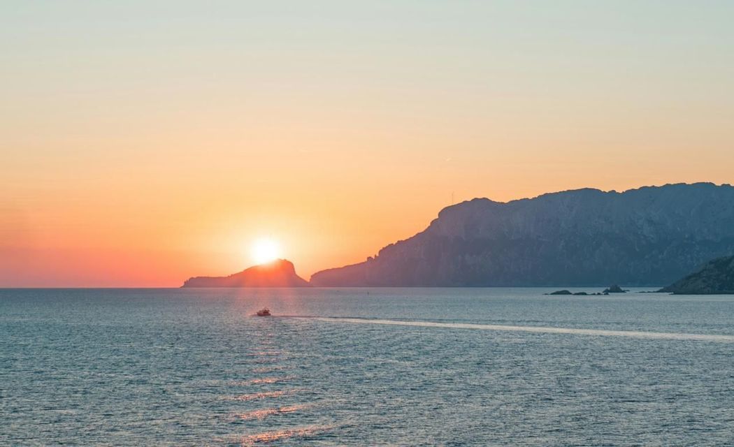 Una piccola barca naviga sul mare mentre il sole tramonta dietro un'isola in silhouette, proiettando un bagliore arancione sull'acqua.