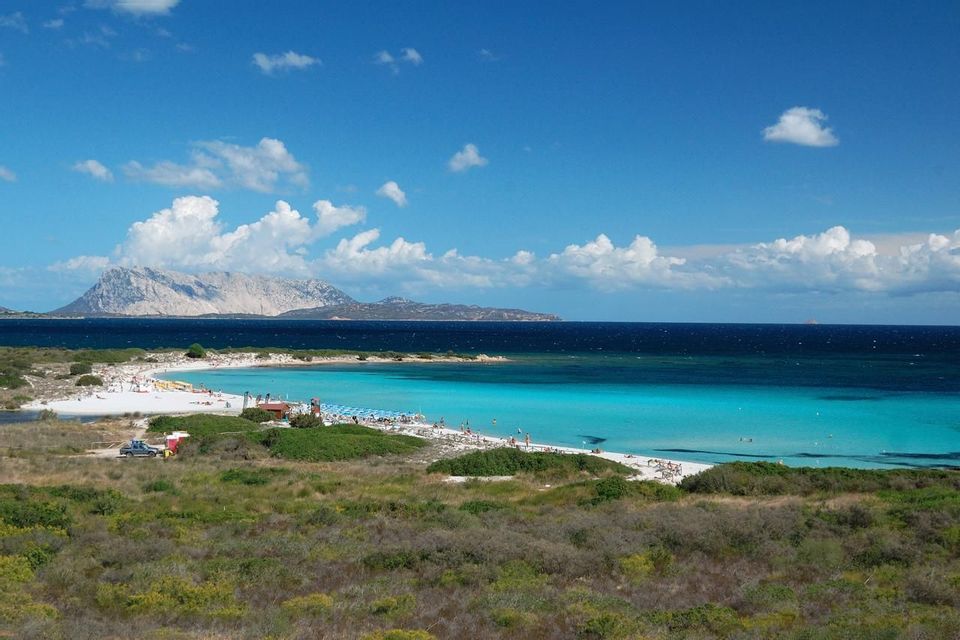 Una vista panoramica di una spiaggia di sabbia bianca e mare turchese, con una grande isola sullo sfondo sotto un cielo parzialmente nuvoloso.