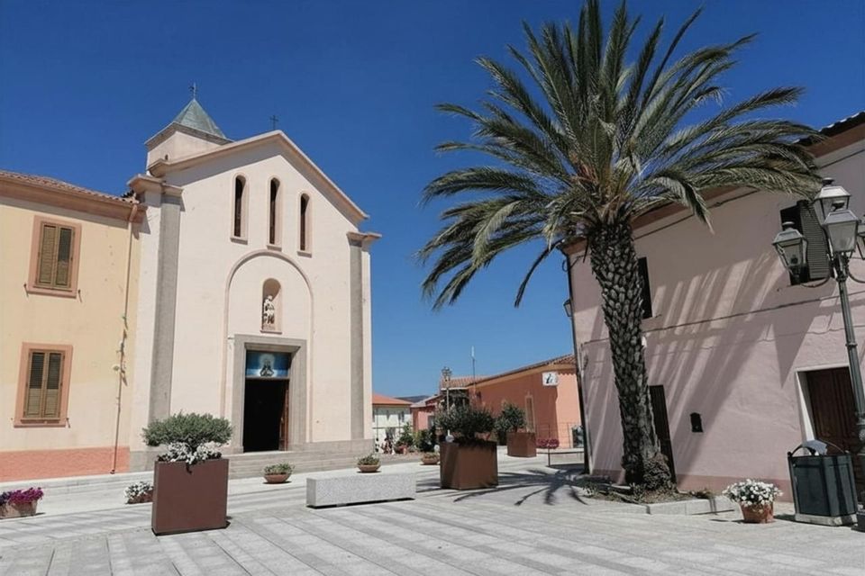 Una chiesa rosa pallido e una grande palma si ergono in una piazza cittadina soleggiata e pavimentata sotto un cielo azzurro e limpido.