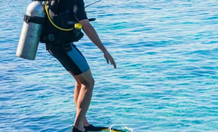 Eine Person in einem Neoprenanzug mit Tauchflasche und Flossen steht bereit, in das klare blaue Wasser einzutauchen.