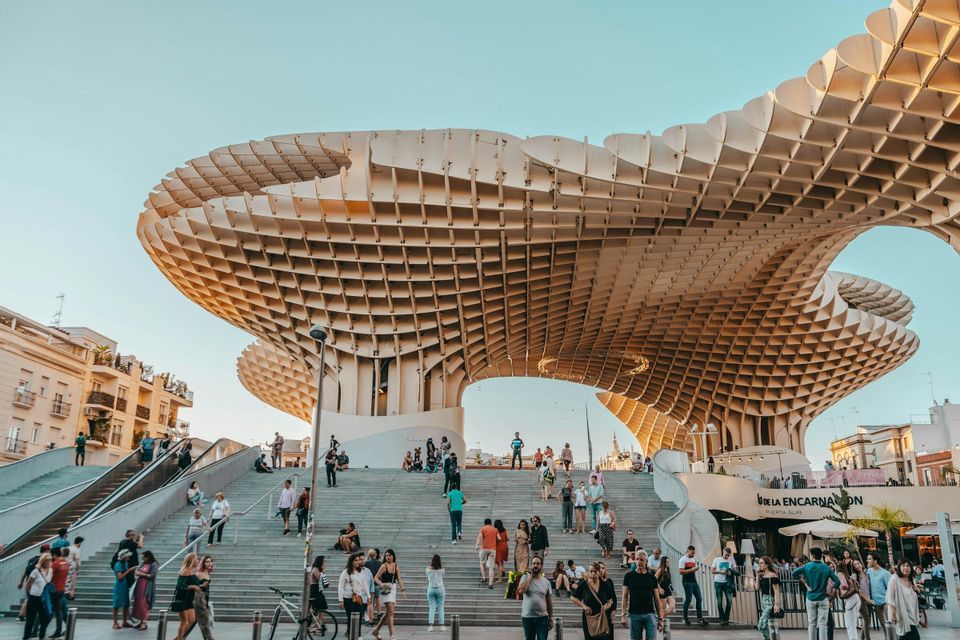 Des personnes marchent et se rassemblent sur de larges escaliers sous un grand auvent architectural moderne en treillis, sous un ciel dégagé.