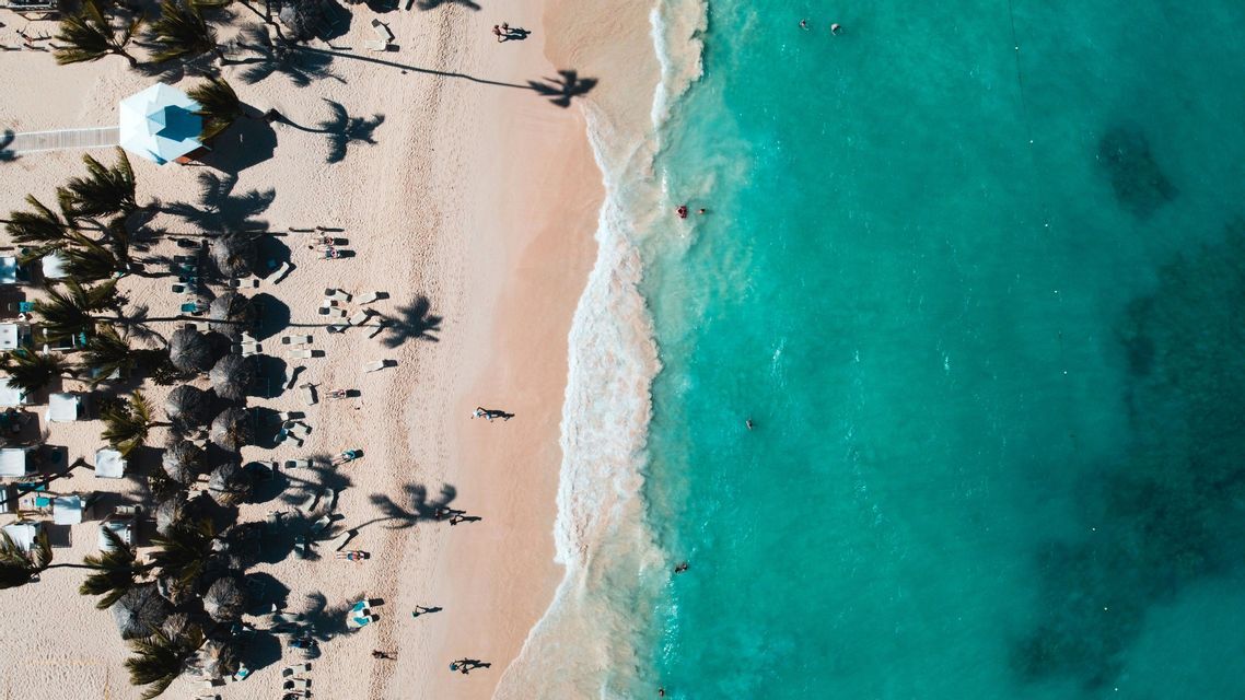 Una vista aerea dall'alto di una spiaggia sabbiosa accanto a un oceano turchese, con persone che nuotano e si rilassano sotto le palme.