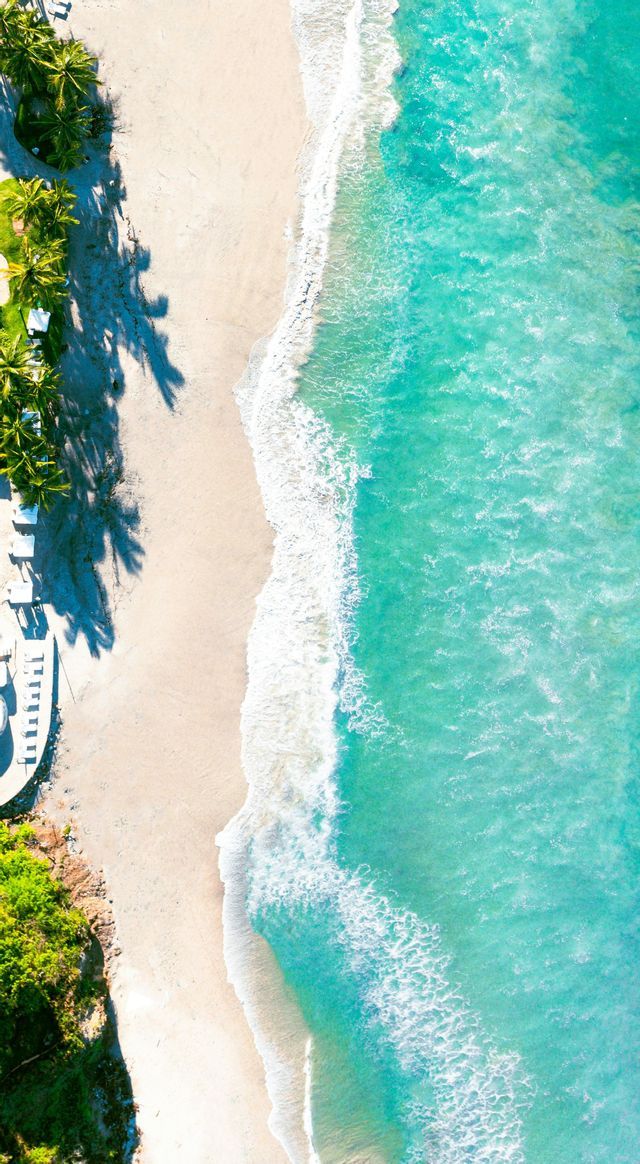 Una vista aerea dall'alto di acqua turchese e onde bianche che incontrano una spiaggia sabbiosa fiancheggiata da palme.