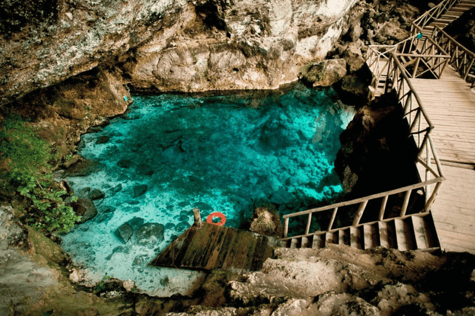 Una scala di legno conduce a un piccolo pontile su un cenote con acqua limpida e turchese all'interno di una grotta rocciosa.