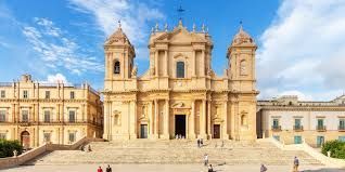 La facciata ornata, color sabbia, di una cattedrale barocca in cima a un'ampia scalinata sotto un cielo blu con nuvole sottili.